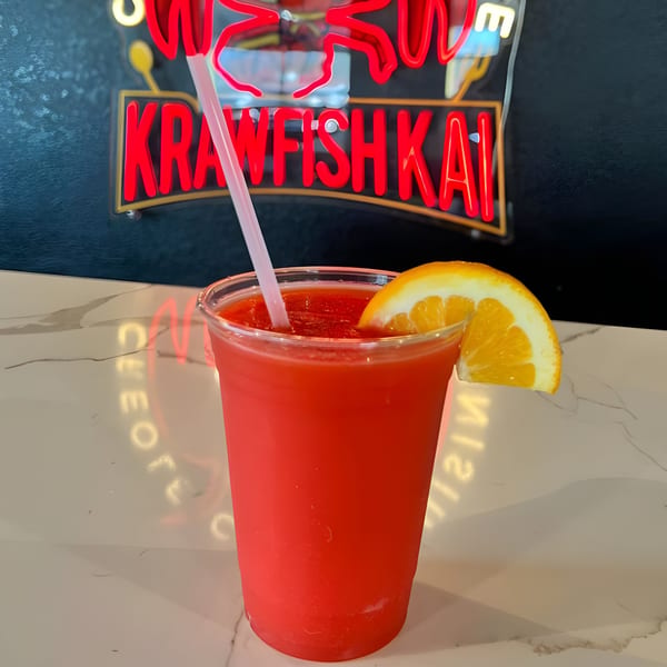 A red frozen drink with an orange slice and straw sits on a counter below a Krawfish Kai neon sign.
